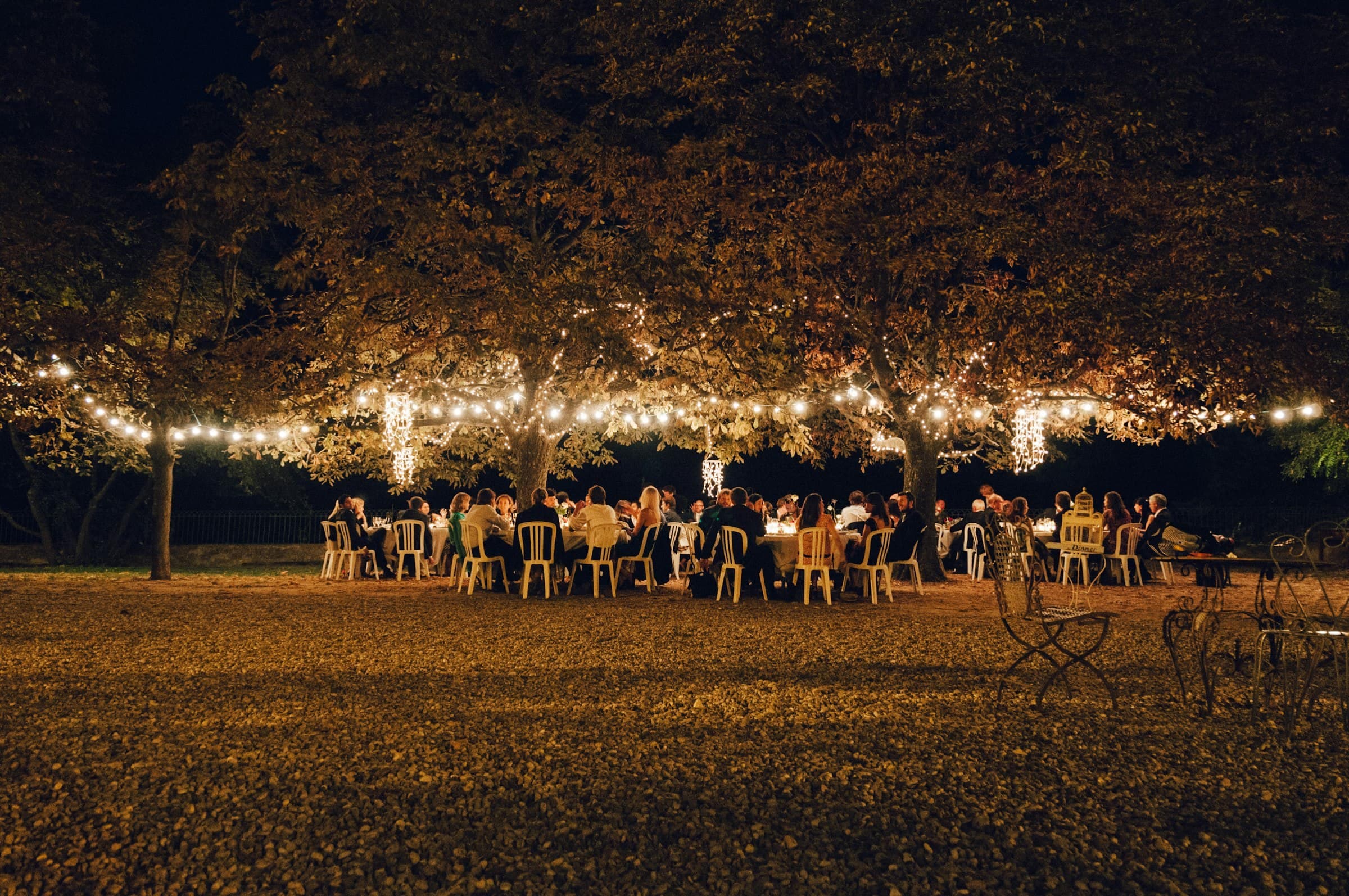 Invitados cenando de noche en un jardín, bajo árboles iluminados con series de luces
